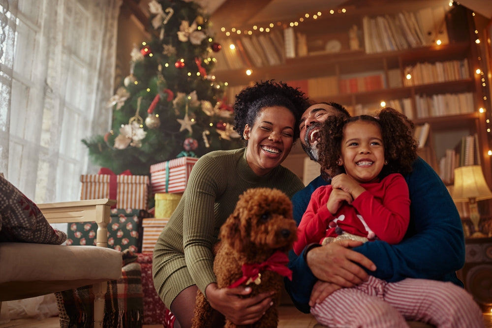 A joyful family, including a woman, a man, a girl in red pajamas, and a dog, laugh together in a cozy living room decorated for the holidays, with a Christmas tree and wrapped gifts in the background.