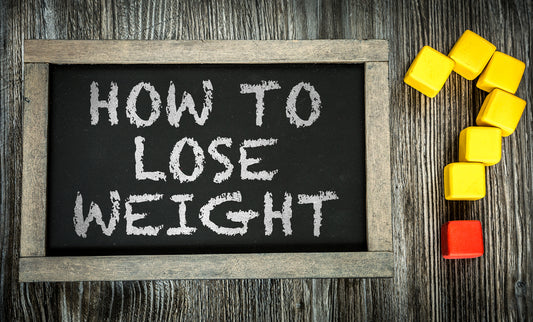 A chalkboard with the phrase "HOW TO LOSE WEIGHT" written in white chalk sits on a wooden surface, accompanied by colorful plastic blocks, including a red block and several yellow ones, arranged in a question mark shape.