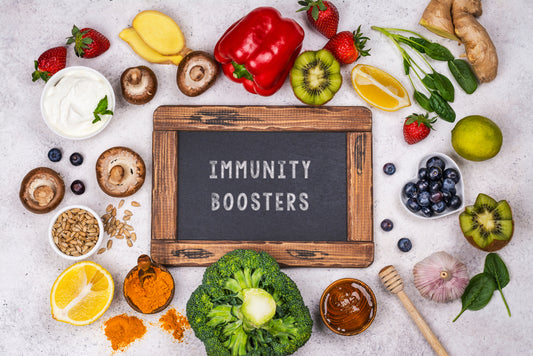 A variety of fresh fruits, vegetables, and spices surround a chalkboard reading "IMMUNITY BOOSTERS," highlighting foods that support immune health.