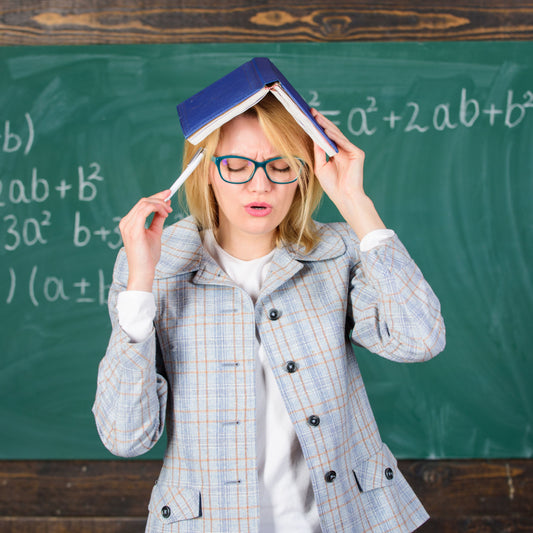 A frustrated woman with blonde hair and glasses holds a book on her head in front of a chalkboard filled with math equations, conveying a struggle with memory or learning concepts.