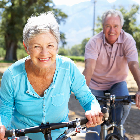 A smiling older woman in a light blue shirt rides a bicycle in a sunny outdoor setting, with a man on a bike behind her, both enjoying a leisurely ride.