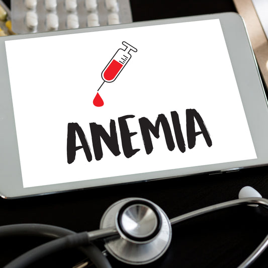 A tablet displays the word "ANEMIA" in bold letters with a graphic of a syringe dripping red liquid, placed on a dark surface next to a stethoscope and a blister pack of pills.