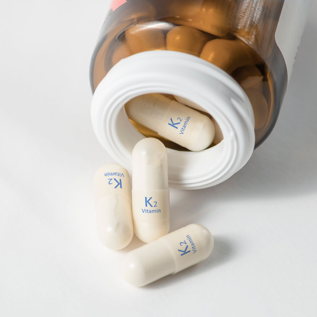 A bottle of vitamin K2 capsules is tipped over, with several white capsules displaying