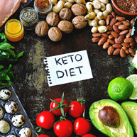 A variety of healthy foods including nuts, tomatoes, avocados, and spices surround a handwritten note that says "KETO DIET," all arranged on a dark surface.