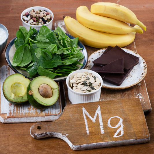 A wooden cutting board with "Mg" written on it is surrounded by bananas, spinach, avocado halves, pumpkin seeds, mixed nuts, and dark chocolate on a rustic wooden table, highlighting magnesium-rich foods.