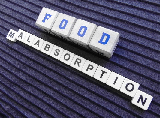 Wooden letter blocks spell "FOOD" above "MALABSORPTION" on a textured purple background, highlighting the relationship between nutrition and nutrient absorption issues.