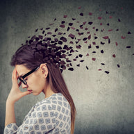 A woman with glasses holds her head in distress, as fragmented shapes appear to disperse from her head against a gray background, symbolizing memory loss potentially linked to vitamin D deficiency.