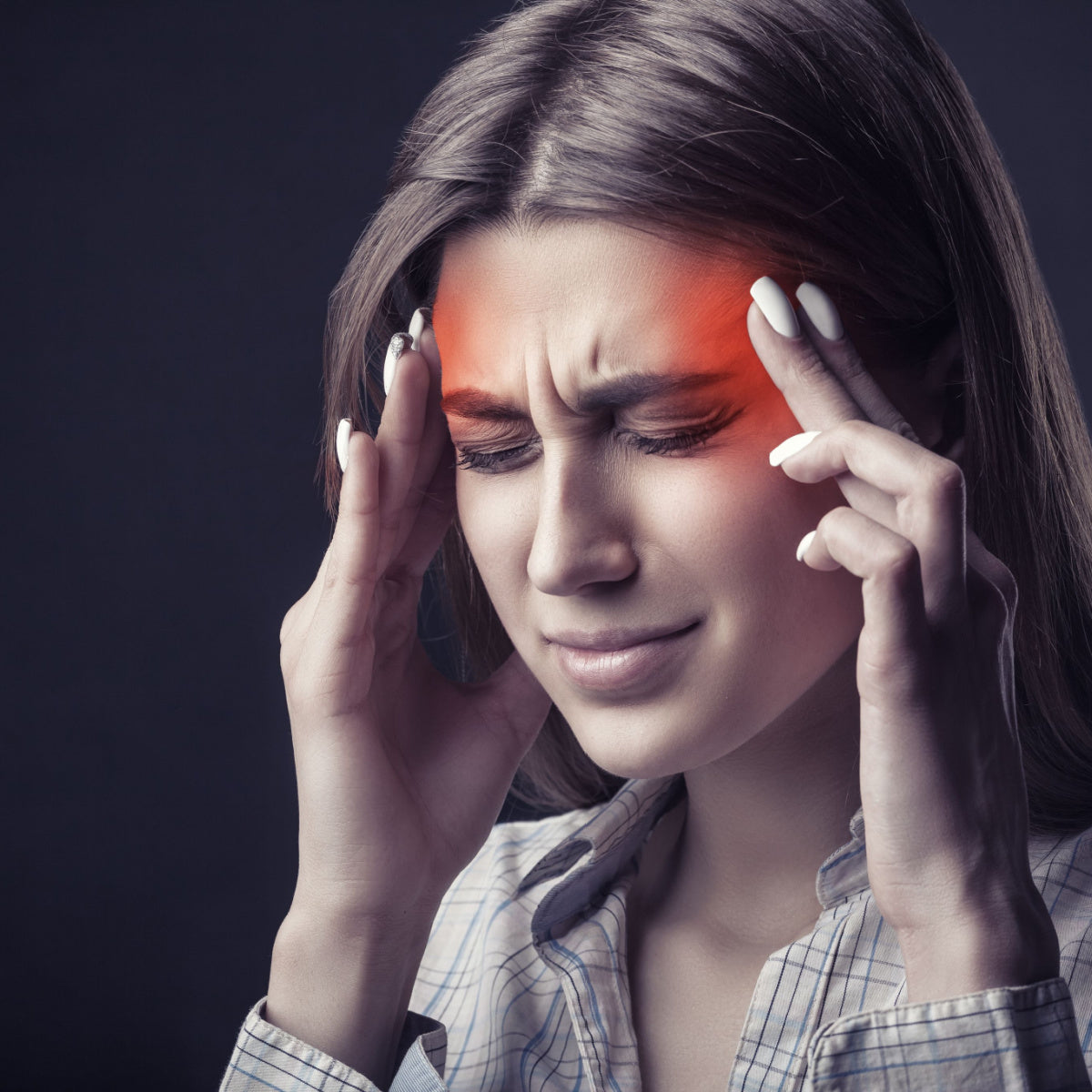 A woman with long hair holds her temples, grimacing in pain, against a dark background, illustrating the discomfort associated with migraines.
