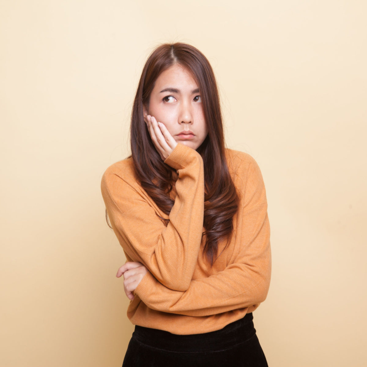 A woman with long hair, wearing an orange sweater, looks thoughtfully to the side while resting her chin on her hand, set against a plain yellow background, conveying a sense of contemplation or concern.