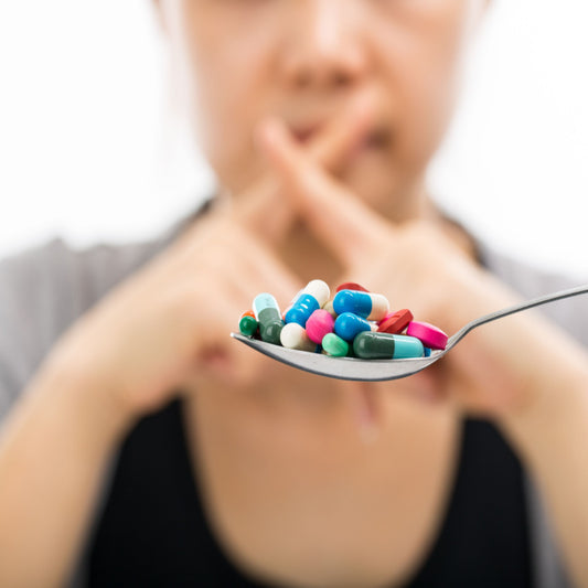 A person in the background makes a gesture of disapproval while a spoon in the foreground holds a variety of colorful vitamins and pills, illustrating a reluctance to take supplements in pill form.