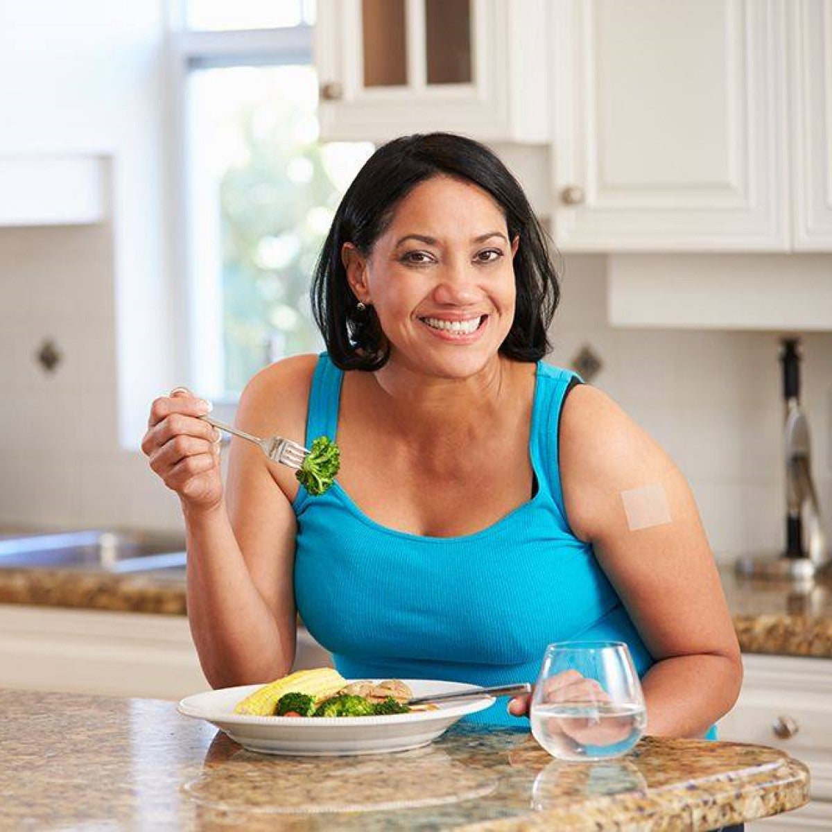 A smiling woman in a blue tank top sits at a kitchen table, holding a fork with broccoli, with a plate of food in front of her and a glass of water beside her, showing a patch on her upper arm.