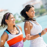 Two women jog along a sunny waterfront, smiling and enjoying their exercise, with buildings and greenery visible in the background.