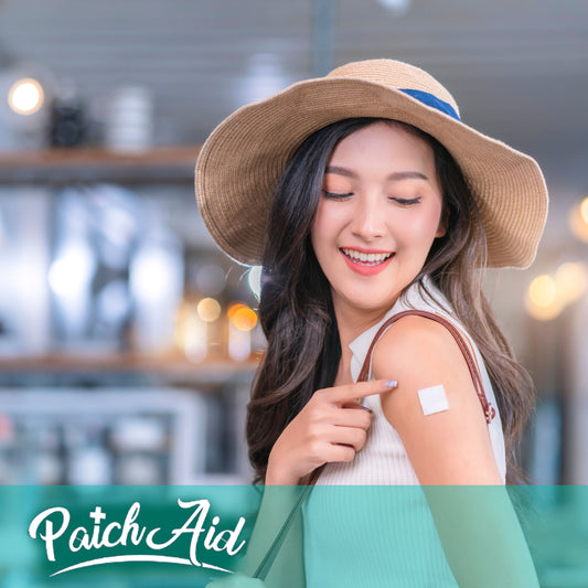 A woman in a wide-brimmed hat smiles and points to a vitamin patch on her arm while standing in a bright, modern setting, highlighting the use of PatchAid's nutritional supplements.