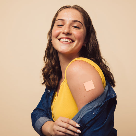 A smiling young woman with wavy hair wears a yellow tank top and denim jacket, showcasing a vitamin patch on her upper arm against a neutral background.
