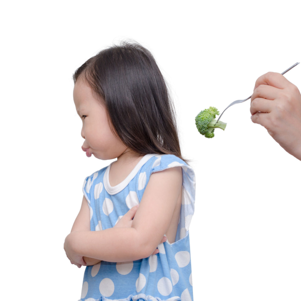 A young girl with long hair and a blue polka-dot shirt makes a displeased face while crossing her arms, as an adult hand offers her a piece of broccoli on a fork.