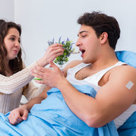 A woman feeds a man sitting in bed, covered with a blue blanket, while a small plant decorates the background, highlighting a caring moment related to his health recovery.