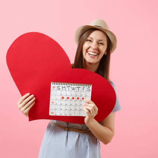A woman in a striped dress and hat smiles while holding a large red heart and a calendar marked with hearts against a pink background.
