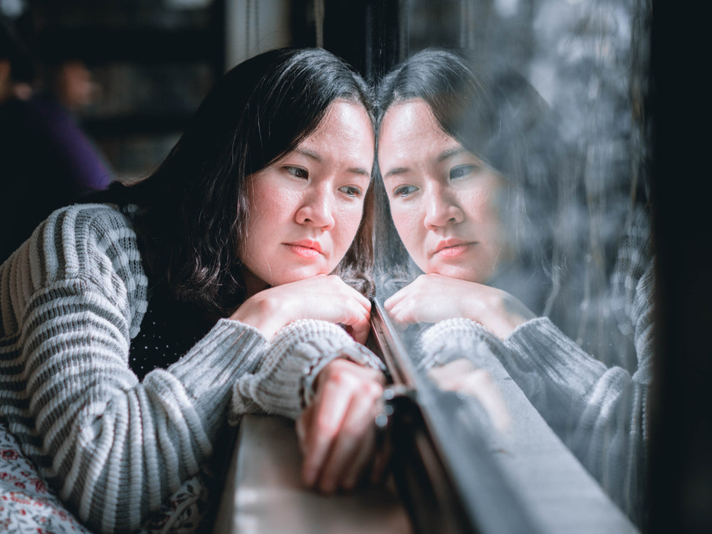 A young woman with long dark hair rests her chin on her hands while gazing thoughtfully out a window, reflecting a sense of contemplation in a cozy indoor setting during winter.