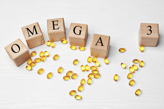 Wooden blocks spelling "OMEGA" and "3" are arranged on a white surface, surrounded by scattered yellow omega-3 gel capsules, highlighting the topic of omega-3 vitamin patches.