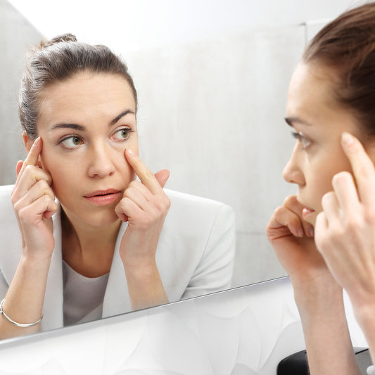 A woman in a white blazer examines her reflection in a mirror, gently pressing her fingers against the skin around her eyes, focusing on reducing signs of aging.
