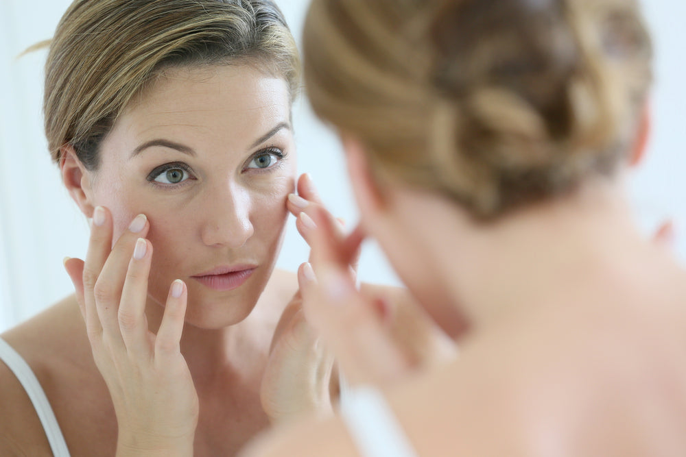 A woman with short brown hair examines her skin closely in a mirror, gently touching her face with her fingers, appearing focused on addressing early signs of aging.