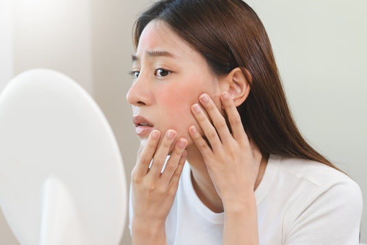 A woman examines her face in a mirror, gently touching her cheek, with a concerned expression, in a bright, neutral-toned room, highlighting her focus on skin health.