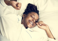 A woman in pajamas smiles and stretches in bed, surrounded by soft white pillows and sheets, conveying a sense of relaxation and restful sleep.