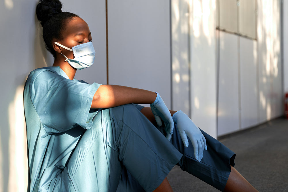 A healthcare worker in scrubs and a mask sits against a wall with eyes closed, appearing exhausted and contemplative, highlighting the toll of sleep deprivation on mental health.