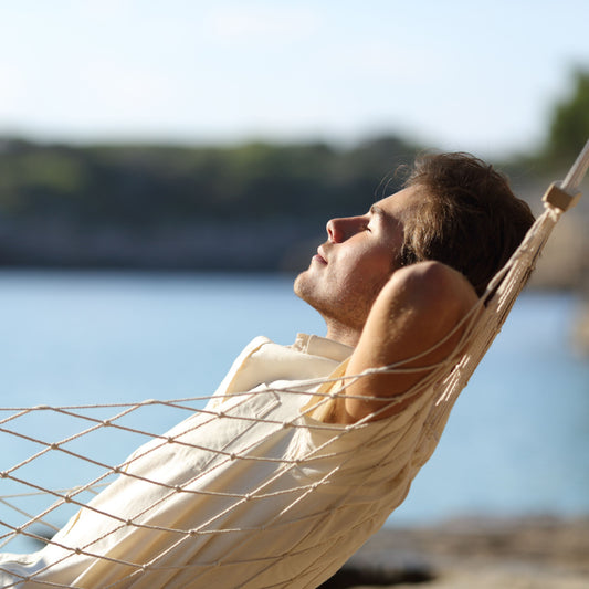 A young man relaxes in a hammock by a calm body of water, eyes closed and face turned upward, enjoying a peaceful moment in a serene outdoor setting.