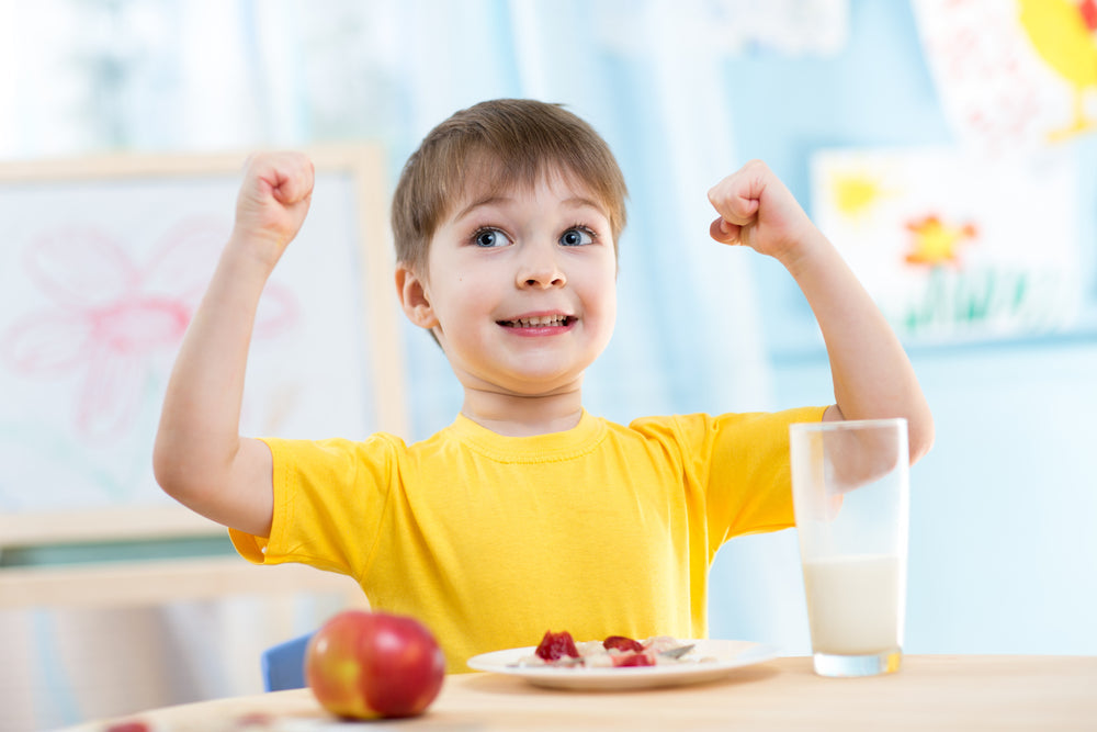 A smiling young boy in a yellow shirt raises his arms in excitement at a table with a plate of food, an apple, and a glass of milk, surrounded by colorful drawings in a bright room.