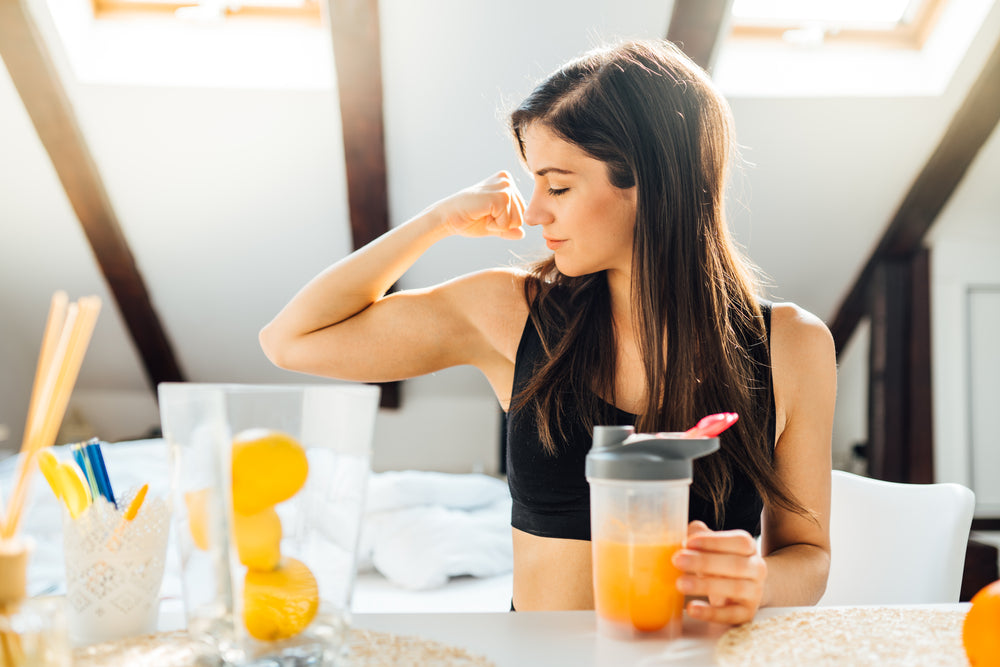 A woman in a black tank top flexes her arm while holding a shaker bottle with a drink, sitting at a table with a glass of lemons and colorful utensils in a bright, airy room.