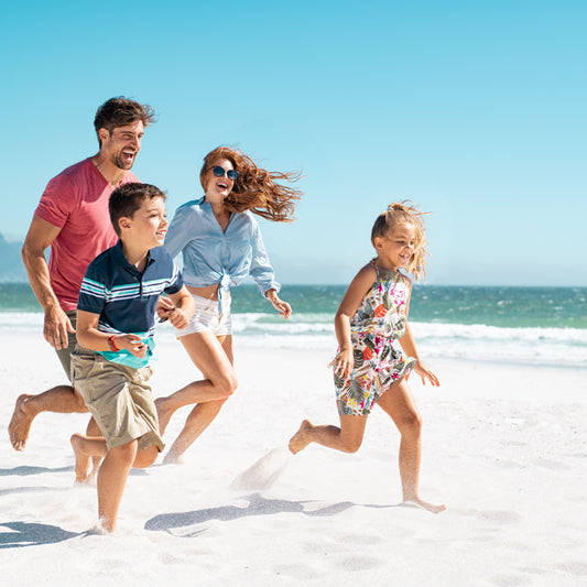 A joyful family of four, including two children, runs barefoot on a sunny beach, laughing and playing together against a backdrop of blue sky and ocean waves.