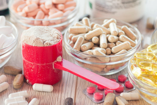 A red measuring scoop filled with powder sits next to various bowls of capsules and tablets, including pink, white, and yellow supplements, arranged on a wooden surface.