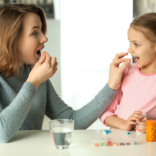 A woman and a girl sit at a table, both smiling as the woman holds a pill while the girl prepares to take one, with a glass of water and a container of pills nearby.