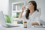 A woman in a white blouse drinks water while sitting at a desk with a laptop, surrounded by vitamin bottles and a notepad, suggesting a focus on health and wellness.