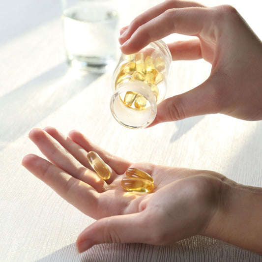 A hand holds a glass jar pouring golden vitamin capsules into an open palm, with a glass of water in the background on a light wooden surface.