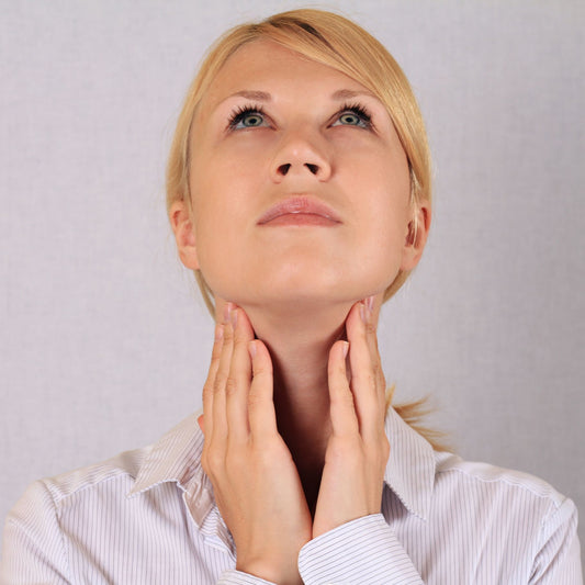 A woman in a striped shirt gently touches her neck while looking upward, suggesting contemplation or concern about thyroid health, set against a plain background.
