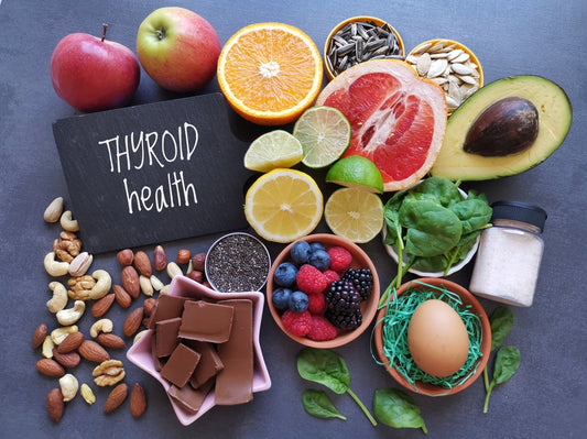 A variety of foods promoting thyroid health, including fruits, nuts, seeds, spinach, an egg, and chocolate, are arranged around a chalkboard sign that reads "THYROID health."