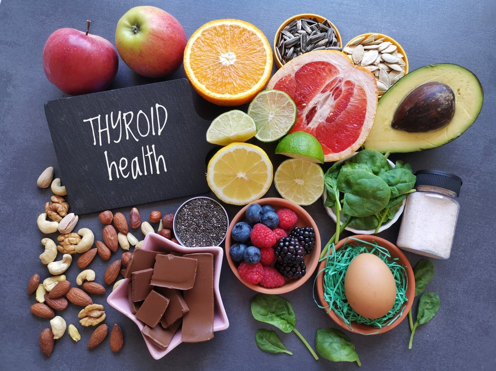 A variety of foods promoting thyroid health, including fruits, nuts, seeds, spinach, an egg, and chocolate, are arranged around a chalkboard sign that reads