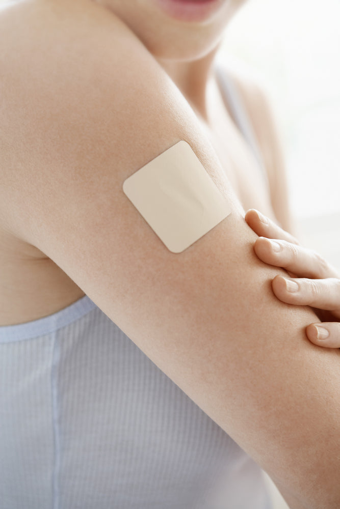 A person is smiling while gently touching a transdermal patch applied to their upper arm, showcasing the patch's placement in a bright, softly lit setting.