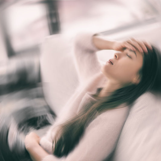 A woman sits on a couch with her eyes closed and hand on her forehead, appearing distressed, suggesting she is experiencing nausea or vertigo in a softly blurred indoor setting.