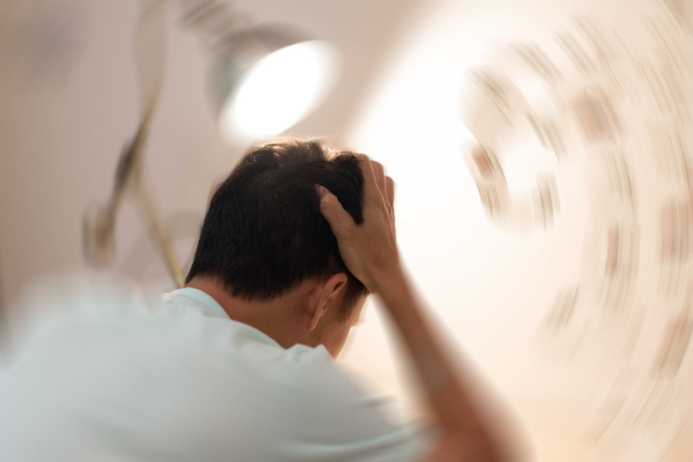 A person sits with their head in their hands, appearing distressed, in a softly lit room, conveying a sense of nausea and vertigo, as blurred lines suggest dizziness around them.