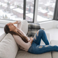 A woman sits on a couch, resting her forehead on her hand with a pained expression, while looking out at a snowy landscape through large windows, suggesting feelings of discomfort or fatigue.