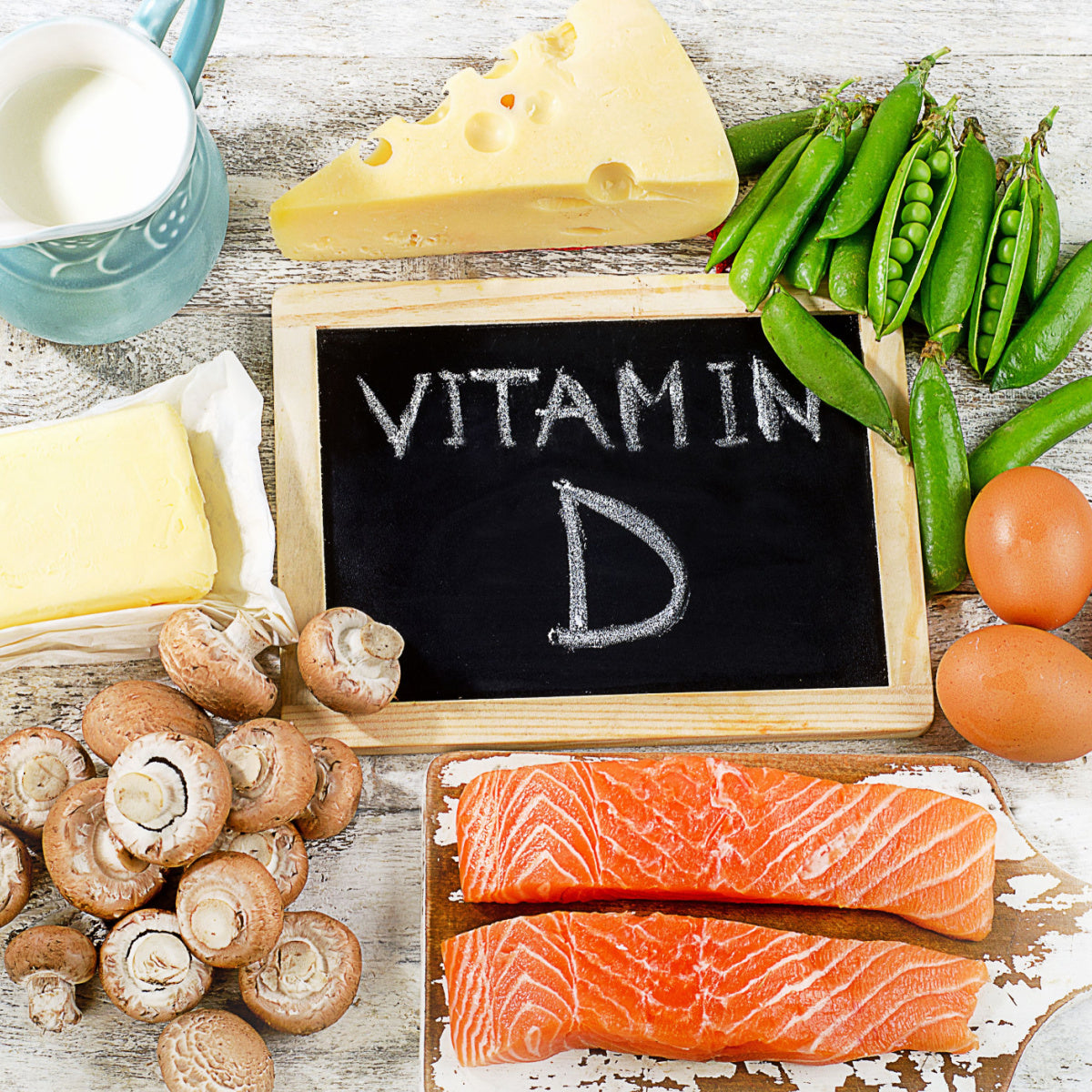 A variety of foods rich in vitamin D, including salmon, eggs, mushrooms, cheese, butter, and green peas, are arranged around a chalkboard displaying