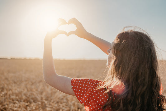 A woman in a polka dot red dress stands in a field, forming a heart shape with her hands around the sun, symbolizing the importance of sunlight for vitamin D and health.