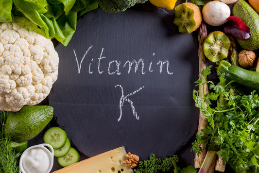 A variety of fresh vegetables and herbs surround a chalkboard with "Vitamin K" written in white chalk, highlighting the importance of this nutrient in a healthy diet.