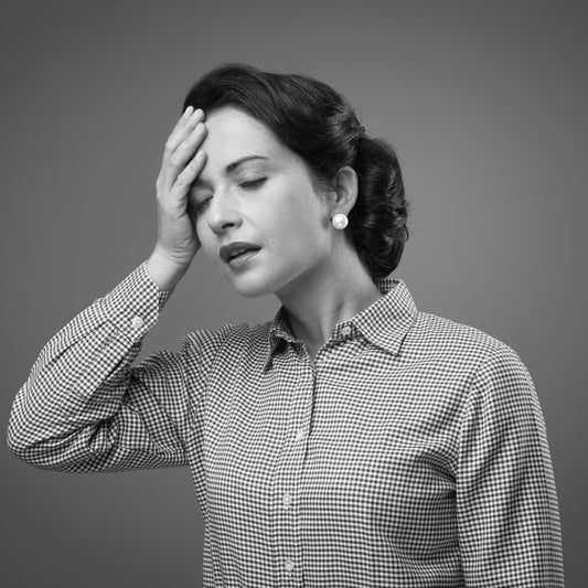 A woman in a checkered shirt holds her forehead with a pained expression, suggesting discomfort or fatigue, set against a plain gray background.