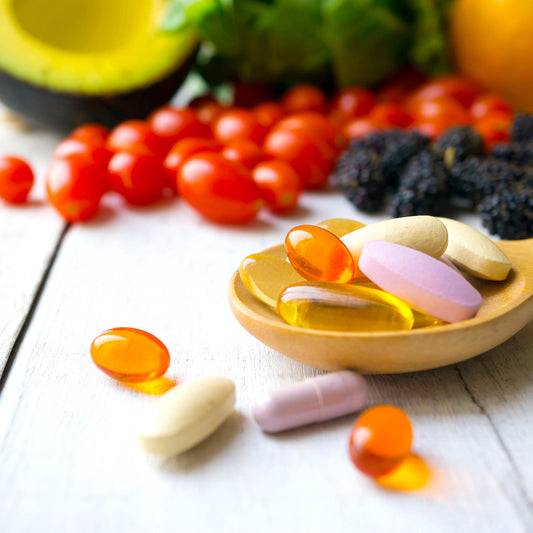 A wooden spoon holds various vitamins and supplements, including colorful gel capsules and tablets, surrounded by fresh vegetables and fruits like cherry tomatoes and avocado on a light wooden surface.
