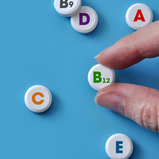 A hand is reaching for a white vitamin tablet labeled "B12" among other tablets marked with letters A, C, D, E, and B9, all set against a light blue background.