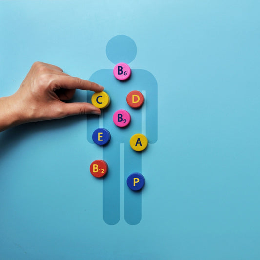 A hand is placing a yellow "C" vitamin capsule on a blue background featuring a silhouette of a person surrounded by colorful vitamin capsules labeled A, B6, B9, B12, D, E, and P.